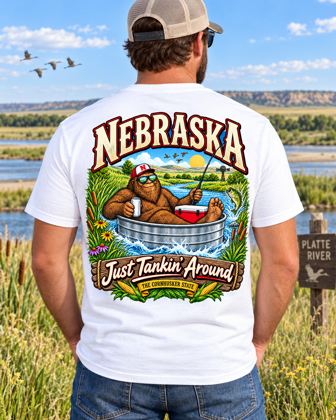 Nebraska Bigfoot shirt with Sasquatch floating in a livestock tank on a river with fishing gear, prairie scenery, corn, and Cornhusker State artwork.